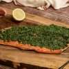 A side of beet green gremolata topped salmon placed on a wooden board, along with a dark colored spoon, half a lemon and two garlic cloves - a white and red kitchen towel is resting in the background