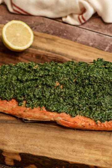 A side of beet green gremolata topped salmon placed on a wooden board, along with a dark colored spoon, half a lemon and two garlic cloves - a white and red kitchen towel is resting in the background