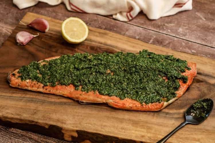A side of beet green gremolata topped salmon placed on a wooden board, along with a dark colored spoon, half a lemon and two garlic cloves - a white and red kitchen towel is resting in the background