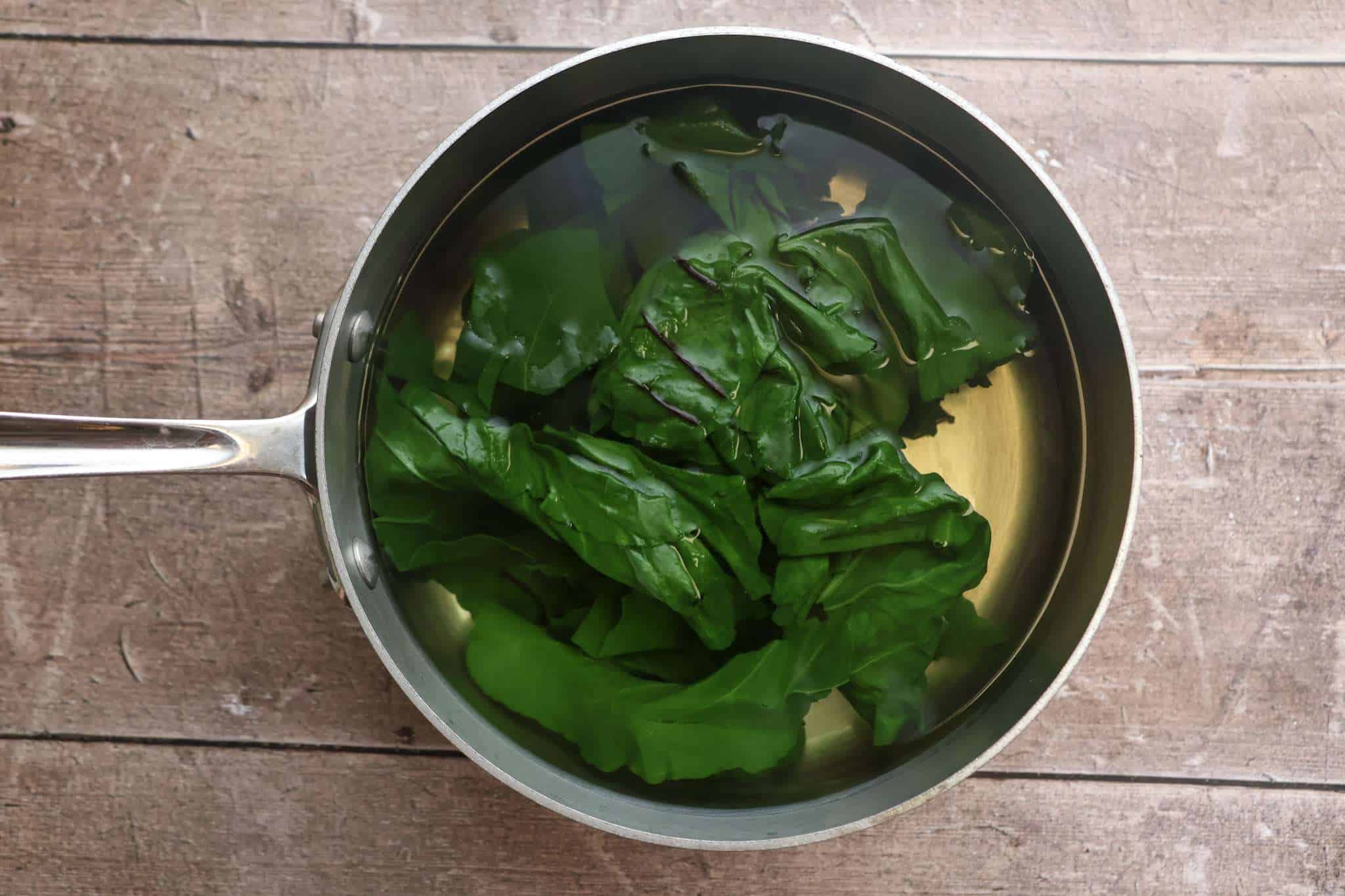A medium stainless steel pot with beet greens that are being cooked/blacnhed