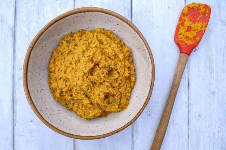 A pottery bowl filled with carrot garlic scape pesto and a spatula resting next to it