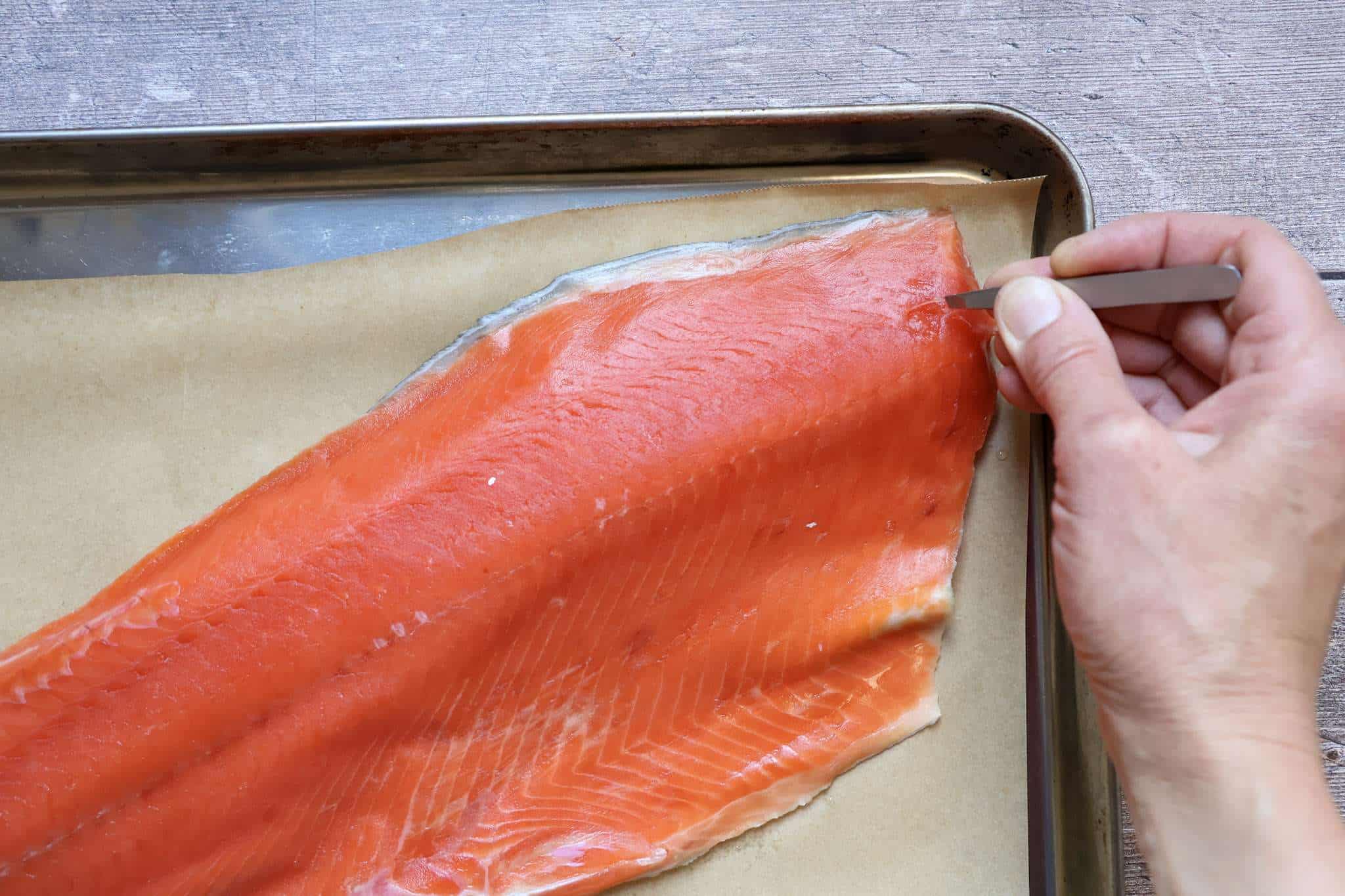 a salmon fillet on a parchment lined baking sheet with a hand holding tweezers on the right hand side of the picture, removing pin bones