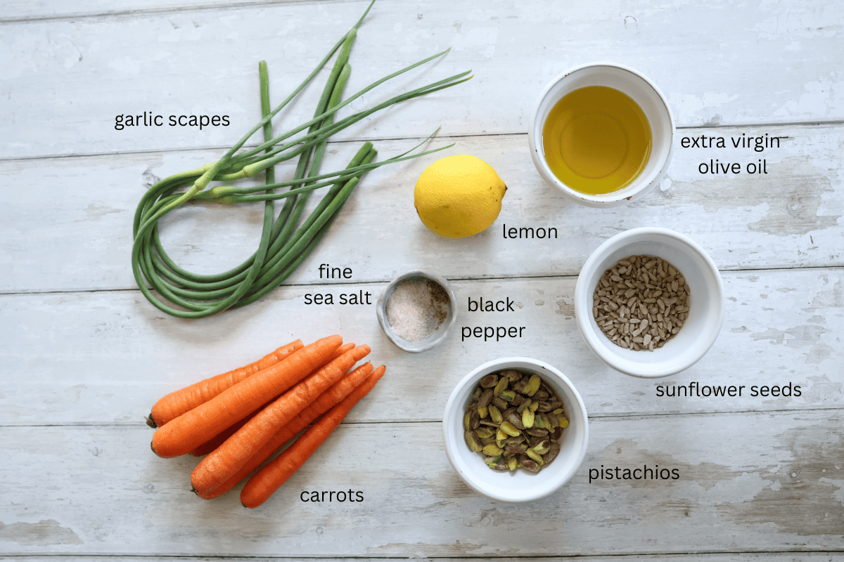 All ingredients for carrot garlic scape pesto are displayed on a white wooden surface and labeled