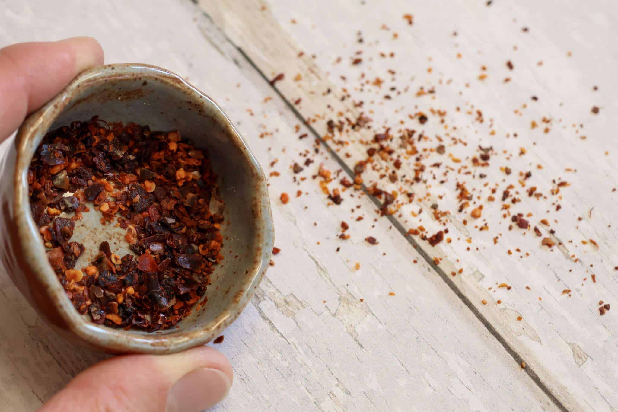A small bowl filled with aleppo pepper and some additional aleppo pepper is spilled out onto the white wooden surface next to the bowl