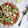 A white bowl filled with cucumber and watermelon salad with feta, mint, lime and aleppo pepper - next to the bowl are a spoon and fork and half a lime