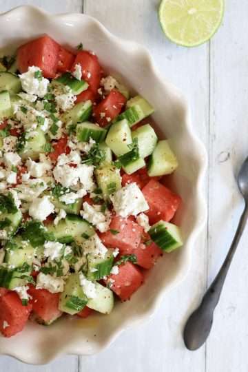A white bowl filled with cucumber and watermelon salad with feta, mint, lime and aleppo pepper - next to the bowl are a spoon and fork and half a lime