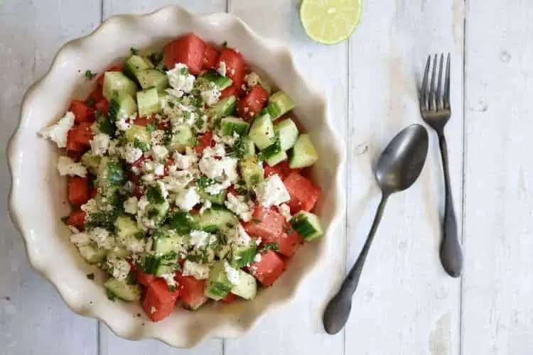A white bowl filled with cucumber and watermelon salad with feta, mint, lime and aleppo pepper - next to the bowl are a spoon and fork and half a lime