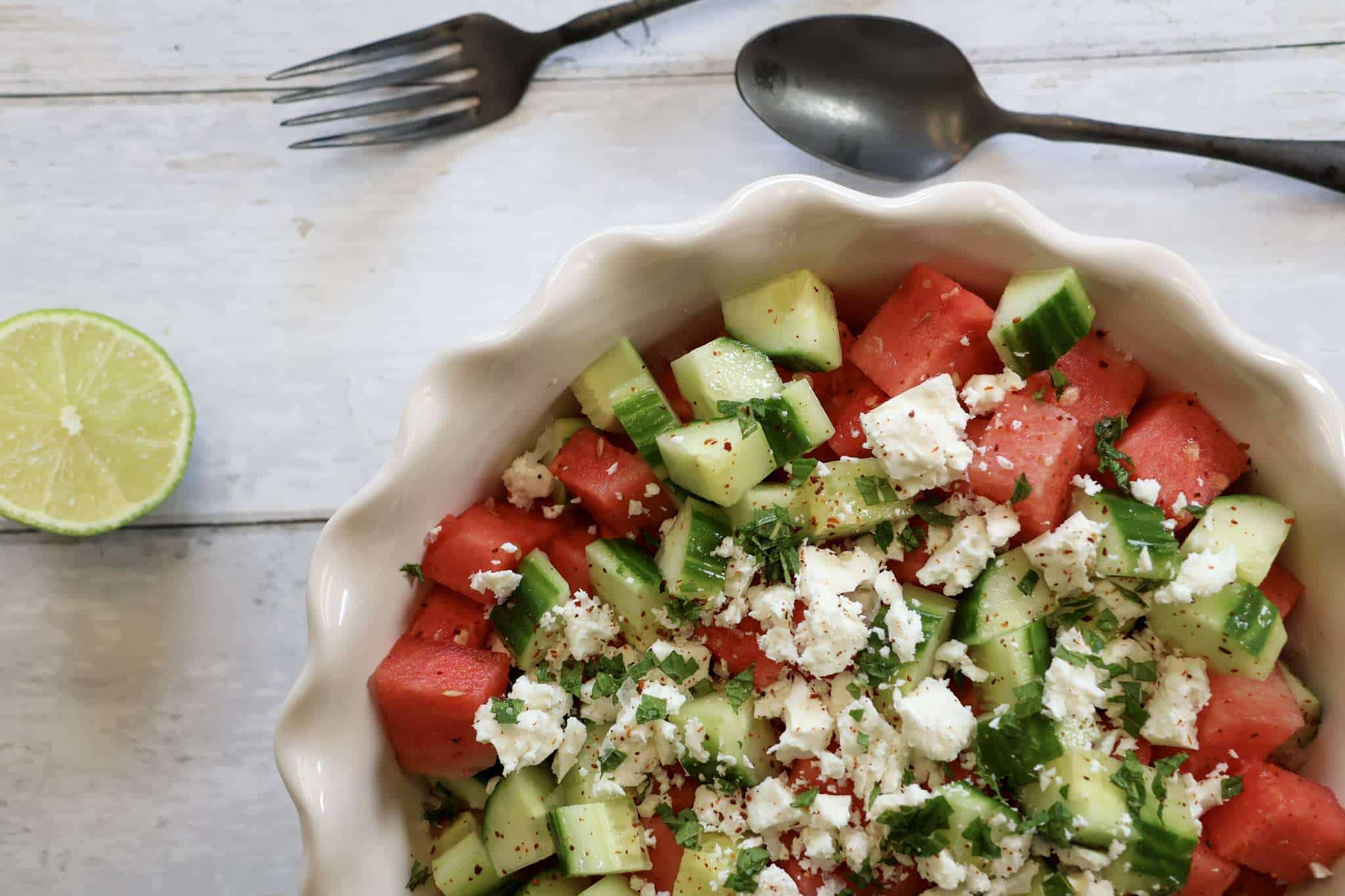 A white bowl filled with cucumber and watermelon salad with feta, mint, lime and aleppo pepper - next to the bowl are a spoon and fork and half a lime