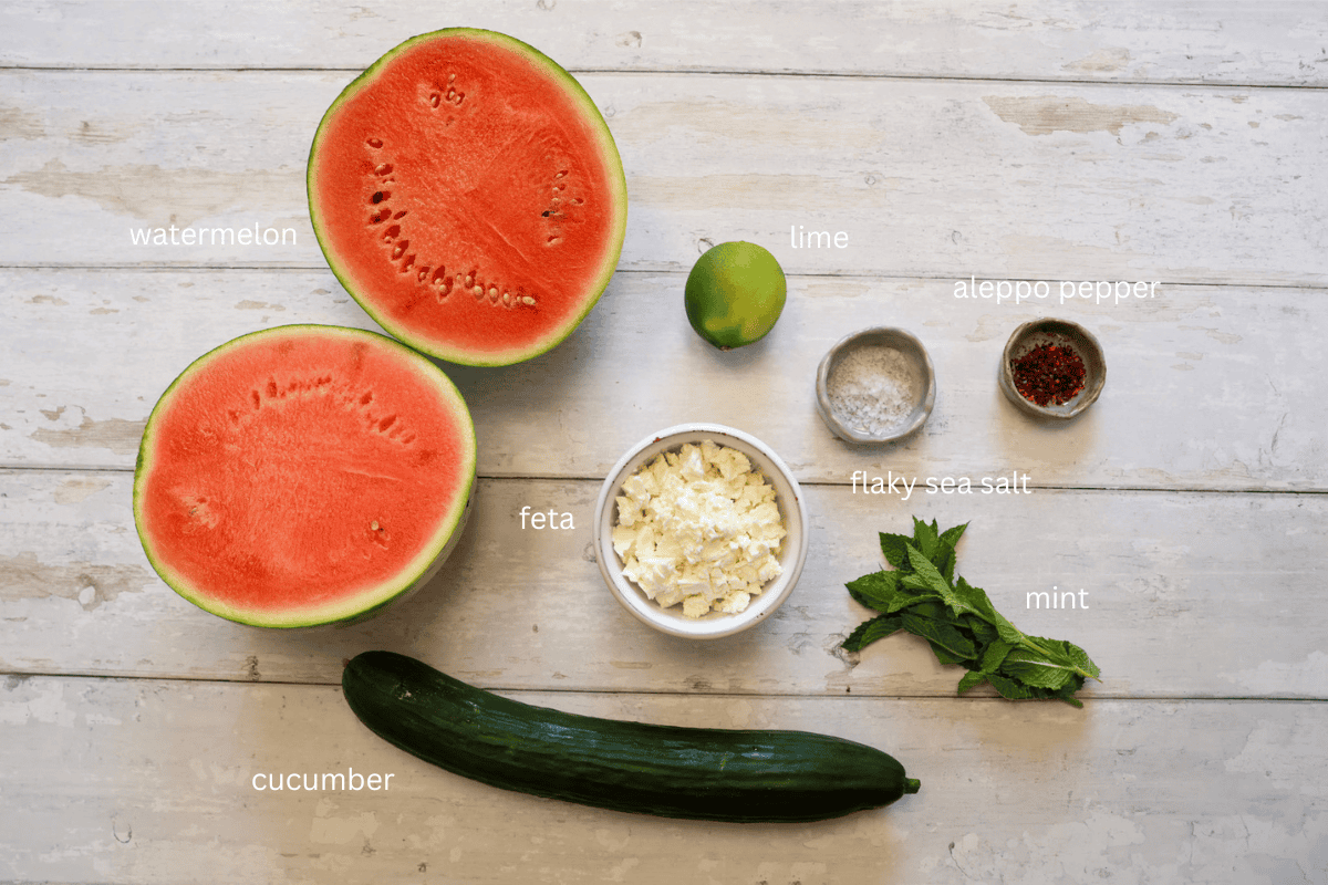 All ingredients for Cucumber and Watermelon Salad with Feta, Mint, and Aleppo Pepper are displayed on a white wooden surface and labeled