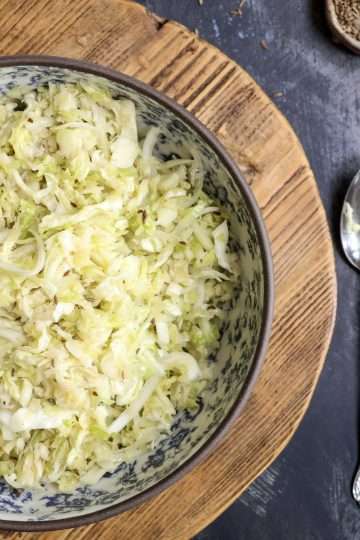 A blue and white floral bowl with German krautsalat is placed on a wooden board and a small bowl of caraway seeds, as well as a fork and spoon are next to the board