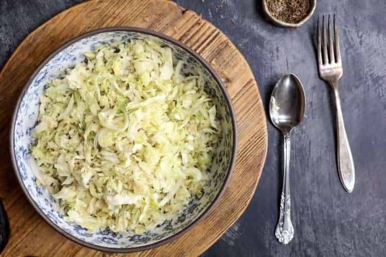 A blue and white floral bowl with German krautsalat is placed on a wooden board and a small bowl of caraway seeds, as well as a fork and spoon are next to the board