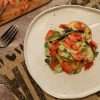 A white pottery plate with smoked salmon, zucchini and peach salad in the center and a few small mint leaves scattered around the plate on the right hand side of the picture - a rustic fork and a small wooden board with additional smoked salmon are partially visible in the picture