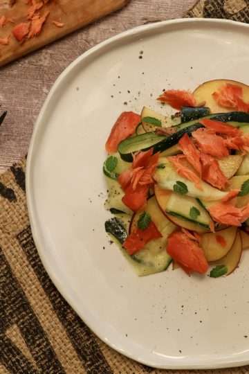 A white pottery plate with smoked salmon, zucchini and peach salad in the center and a few small mint leaves scattered around the plate on the right hand side of the picture - a rustic fork and a small wooden board with additional smoked salmon are partially visible in the picture