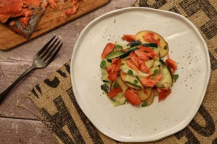 A white pottery plate with smoked salmon, zucchini and peach salad in the center and a few small mint leaves scattered around the plate on the right hand side of the picture - a rustic fork and a small wooden board with additional smoked salmon are partially visible in the picture