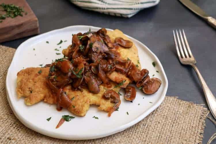 A white plate with mushroom topped chicken schnitzel sprinkled with parsley - a fork, a knife, a beige and green striped napkin and a wooden board with additional parsley can be seen in the background