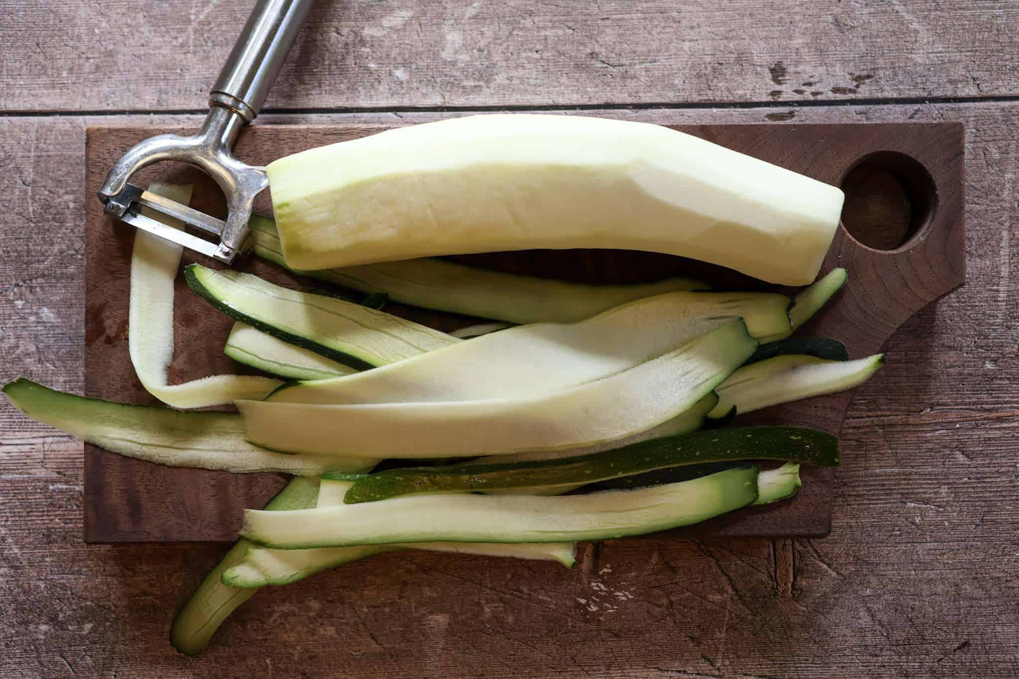 A wooden board with a whole zucchini that has been partially sliced with a vegetable peeler - the peeler is resting on the side of the board