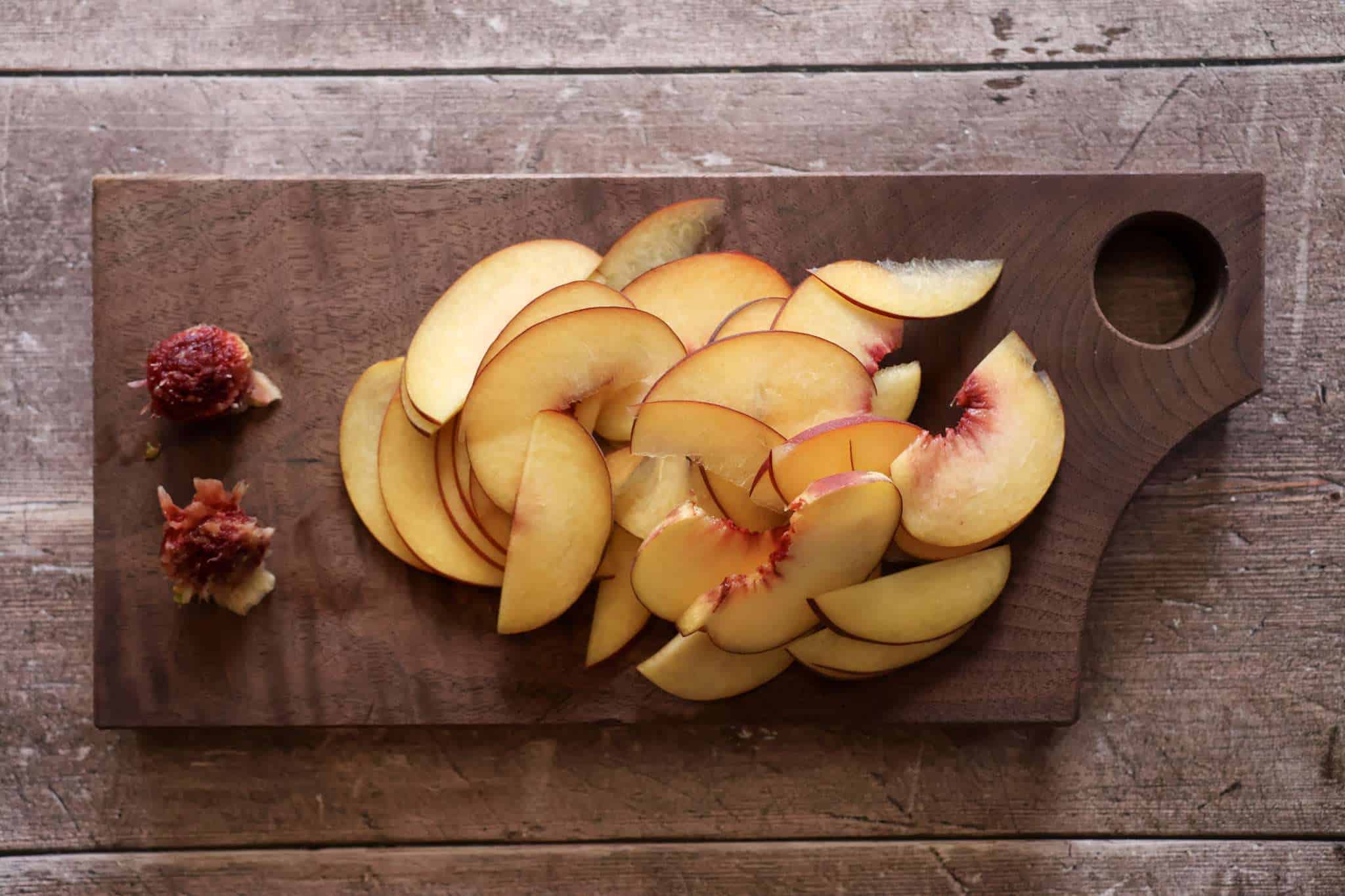A wooden board with thinly sliced peaches and two peach pits