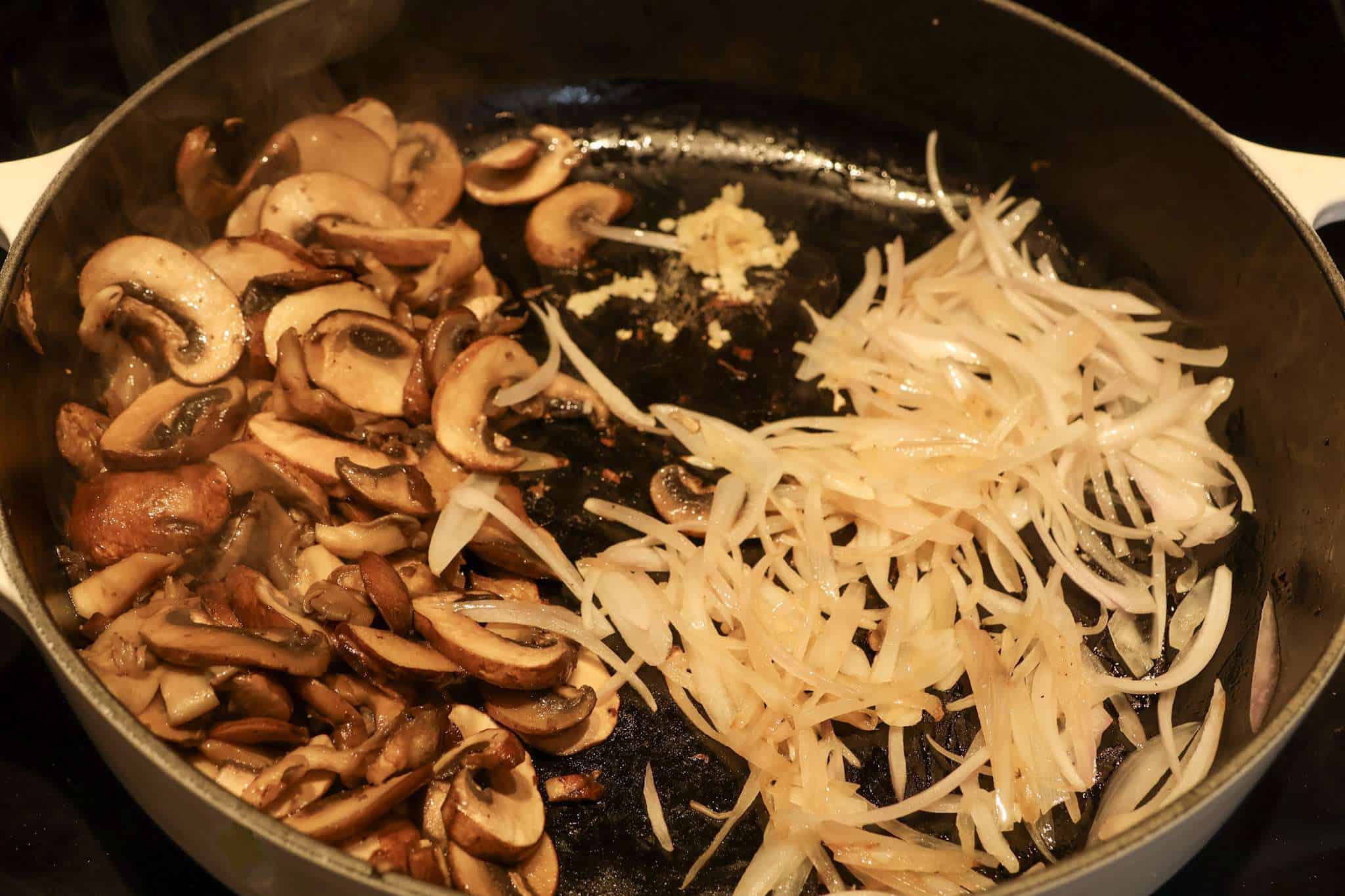 A white cast iron enameled pan with sauteed mushrooms on the left side and sauteed shallots on the right