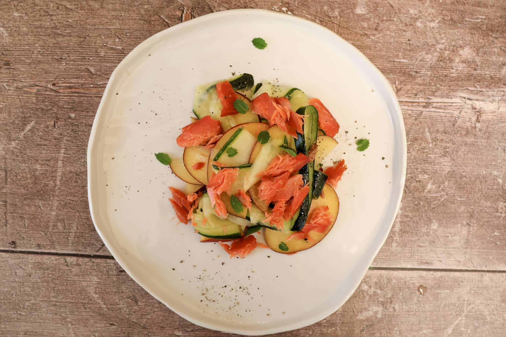 A white pottery plate with smoked salmon, zucchini and peach salad in the center and a few small mint leaves scattered around the plate