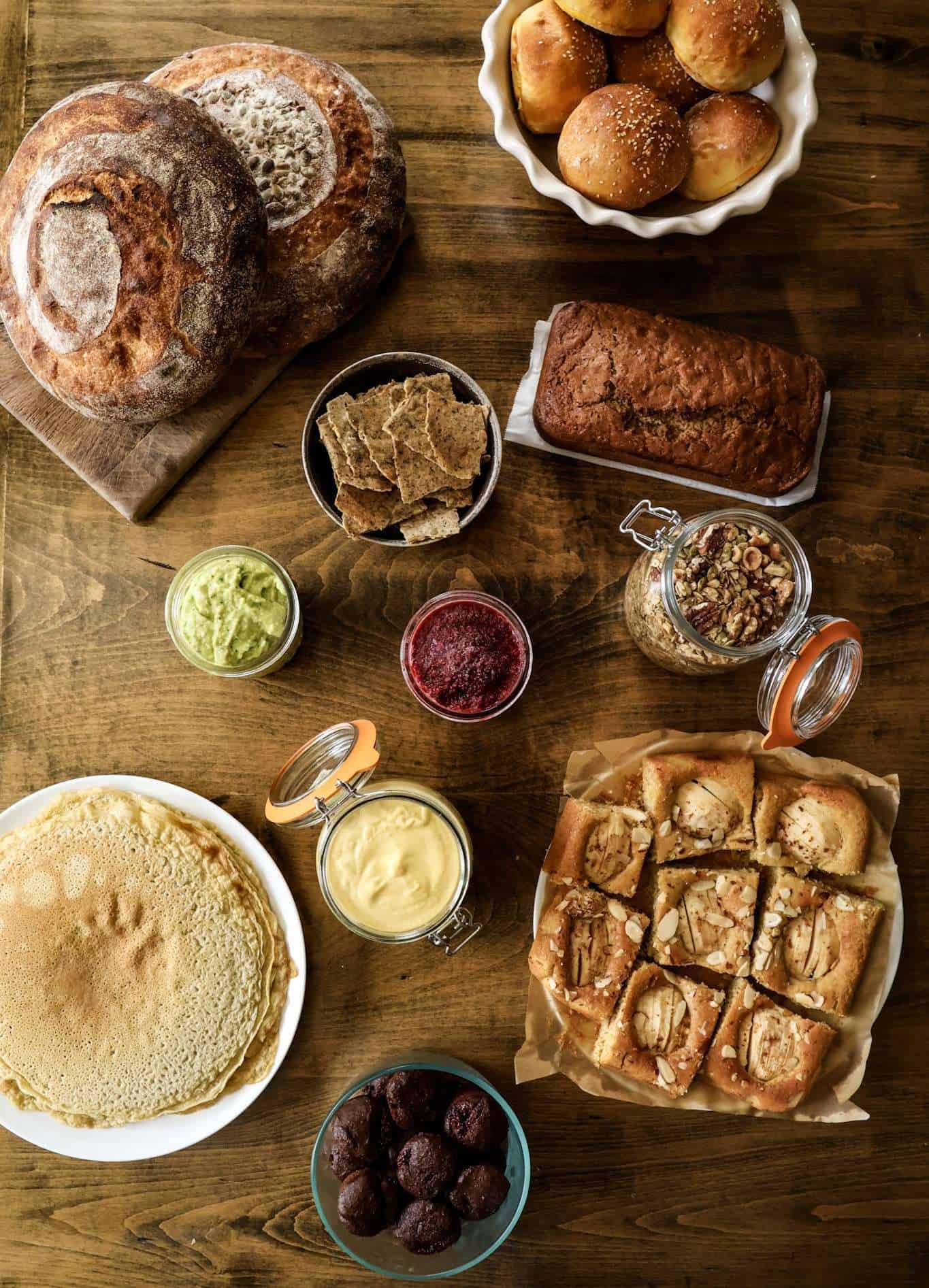 A variety of foods - sourdough bread, rolls, apple cake, granola, hummus, chia jam, crackers, pesto, crepes - displayed on a table