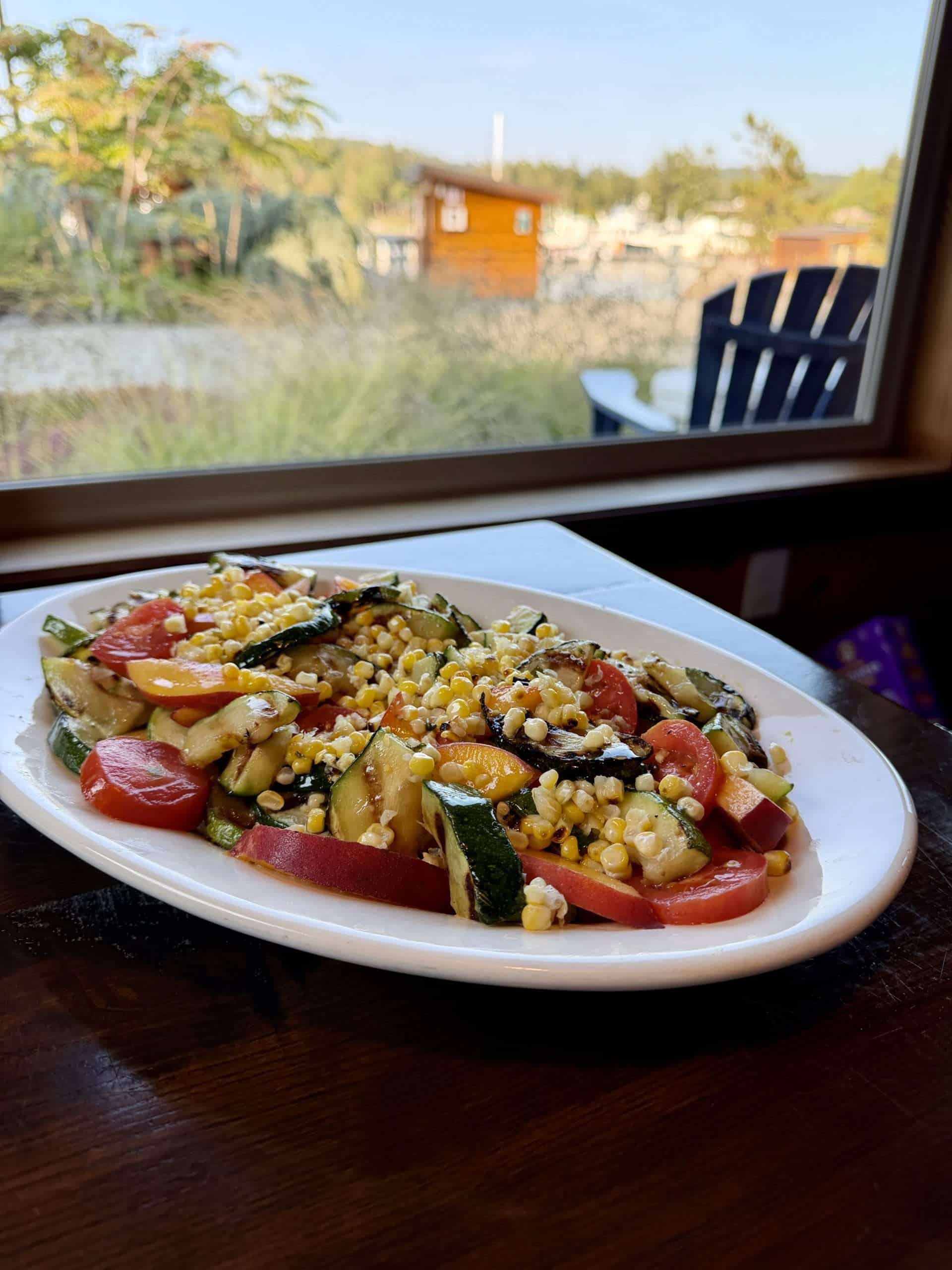 A salad of grilled corn, zucchini, tomatoes, and peaches on a white oval platter on a table right by a window overlooking a small harbor
