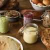 A variety of foods - sourdough bread, rolls, apple cake, granola, hummus, chia jam, crackers, pesto, crepes - displayed on a table