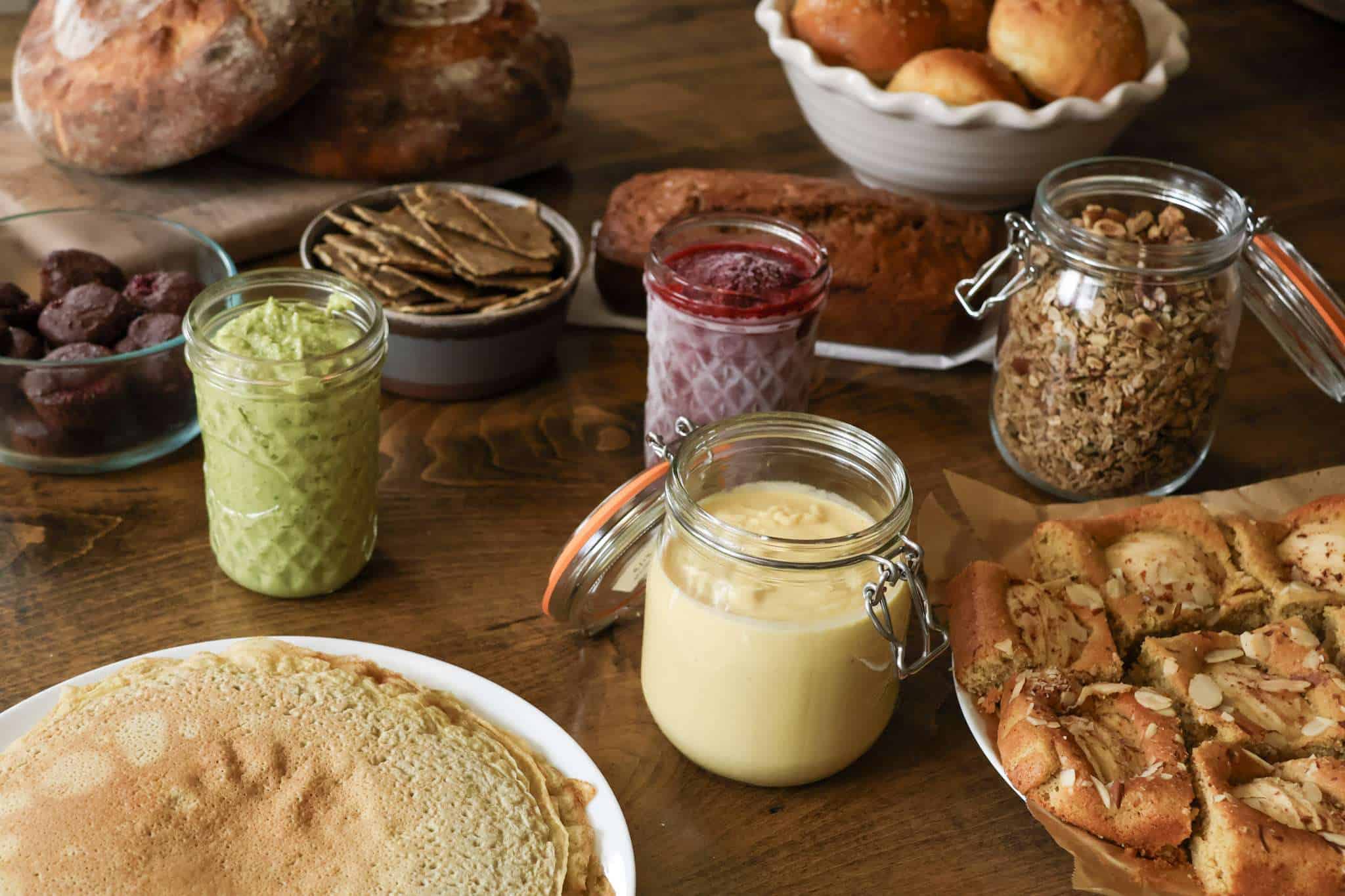 A variety of foods - sourdough bread, rolls, apple cake, granola, hummus, chia jam, crackers, pesto, crepes - displayed on a table
