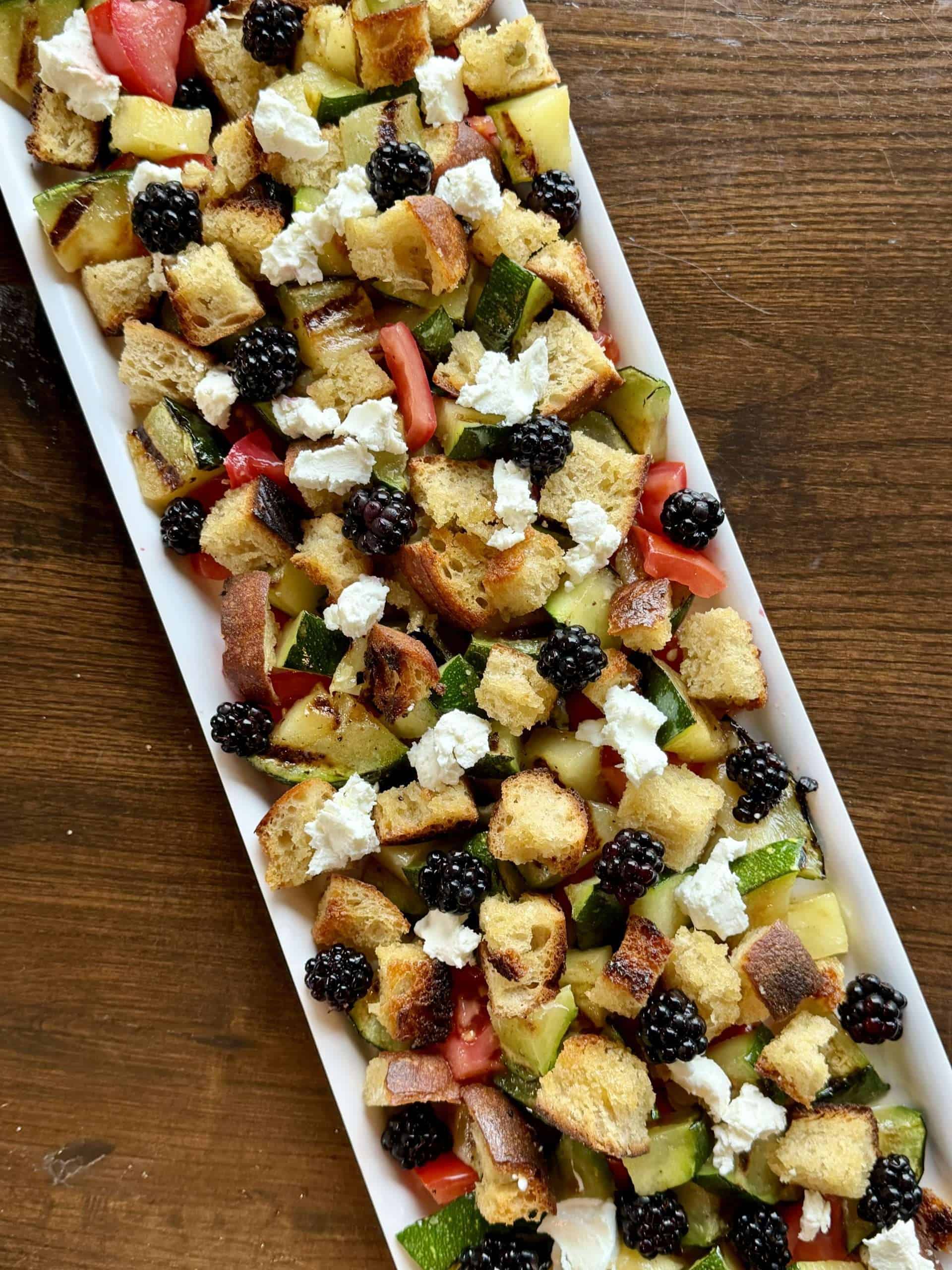 A panzanella salad with blackberries, tomatoes, zucchini, and goat cheese on a white rectangular serving dish
