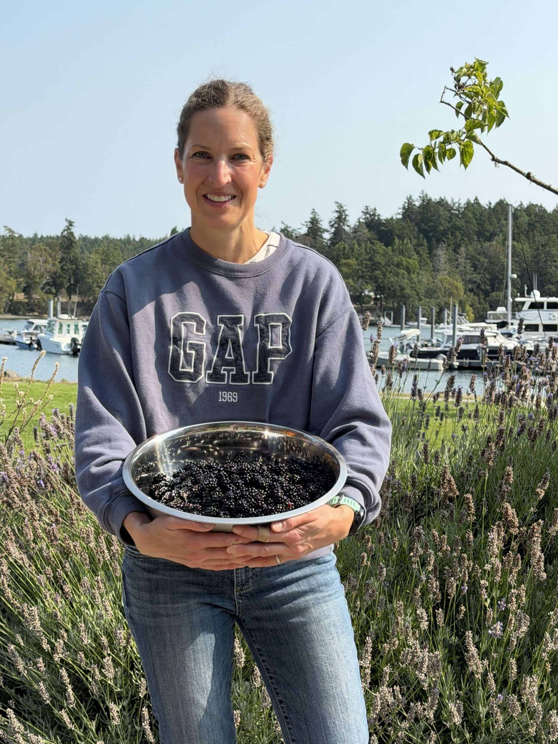 A woman holding a large bowl full of blackberries with a small harbor in the background