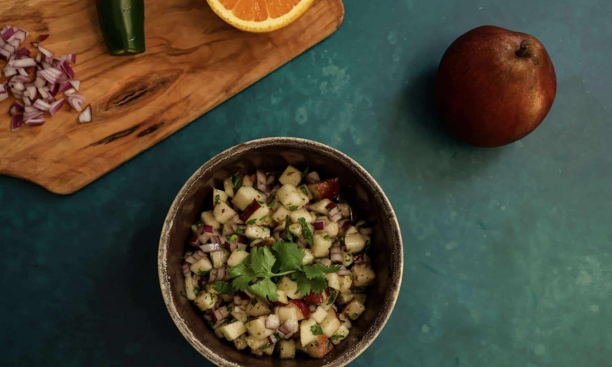 a bowl of pear salsa along with a whole pear and a cutting board holding half an orange, a jalapeno pepper and diced red onions