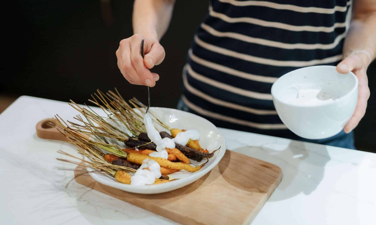 a bowl of roasted carrots with green tops is sitting on a wooden board and a person can be partially seen, drizzling sauce with a spoon with one hand and holding a small white bowl with the other hand
